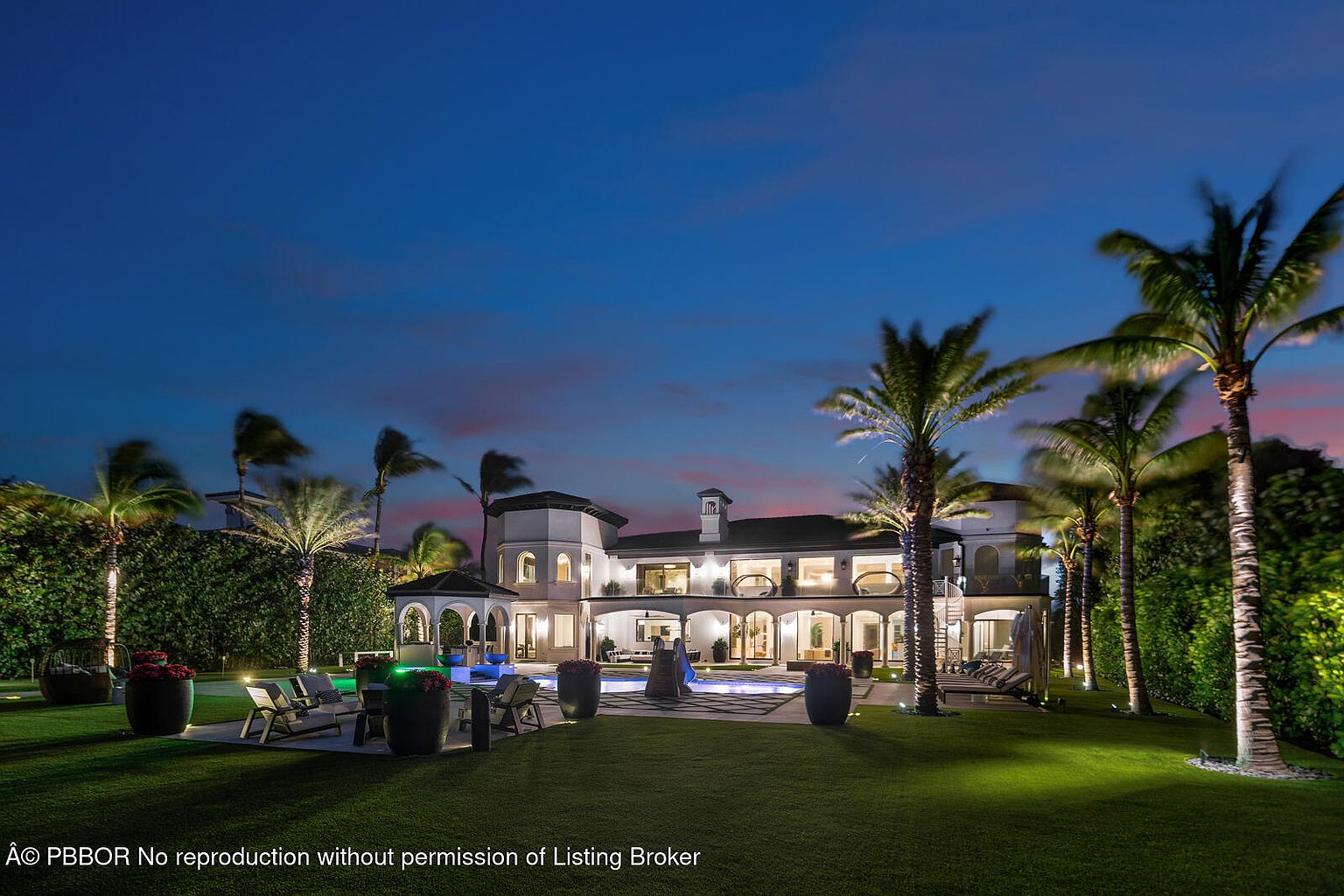 This image showcases the rear exterior of a luxurious home at dusk, featuring a well-manicured lawn, a swimming pool, and several palm trees. The house is illuminated, highlighting its architectural details, including arched windows and a tower-like structure. The scene evokes a sense of opulence and tranquility, perfect for upscale living.