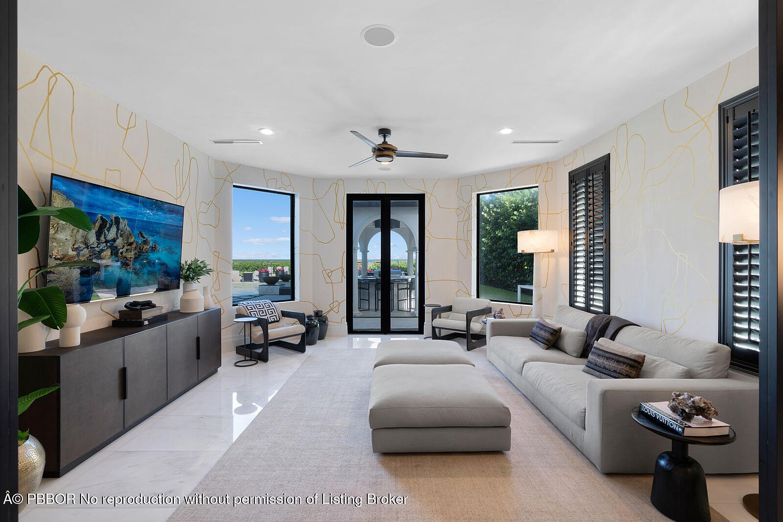 This is an interior shot of a modern living room featuring a neutral color palette with gold accents. The room is well-lit with natural light coming through large windows and features a large sectional sofa, ottoman, and a dark wood media console with a large television. The walls are decorated with a unique gold line pattern, and the flooring appears to be light-colored marble or tile, creating a luxurious and inviting atmosphere.