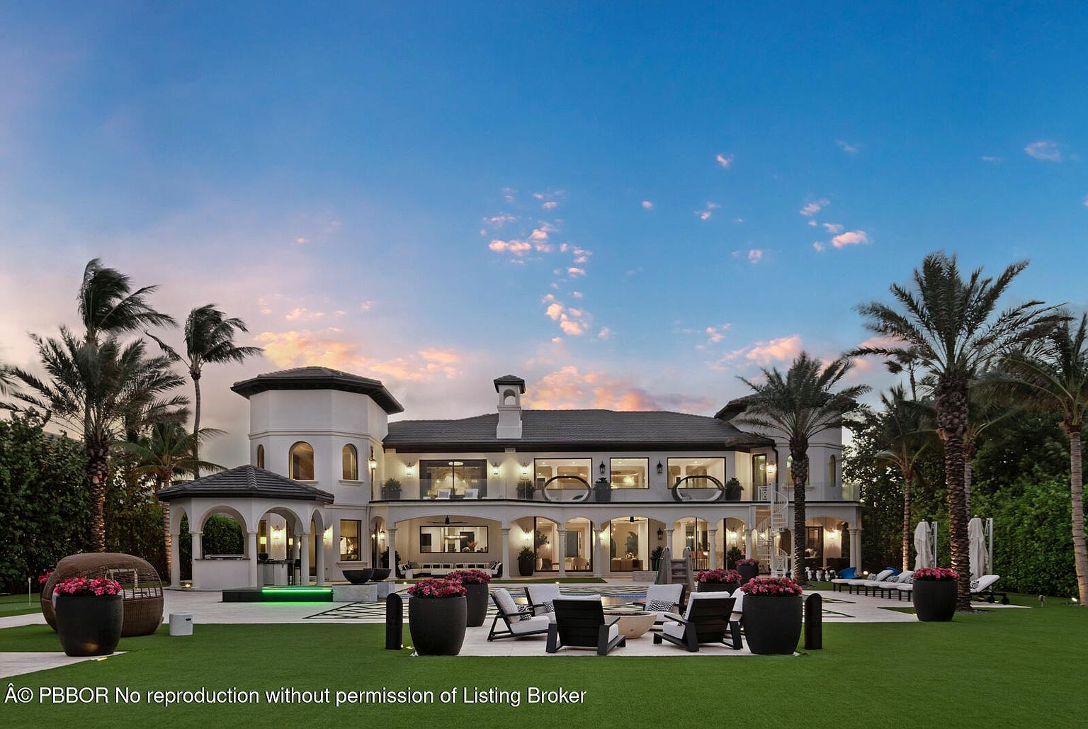 This is a rear view of a luxurious two-story home with a modern design. The property features a well-manicured lawn, a patio area with outdoor seating, and palm trees surrounding the perimeter. The architecture includes arched doorways, balconies, and a central chimney, creating an elegant and inviting outdoor living space.