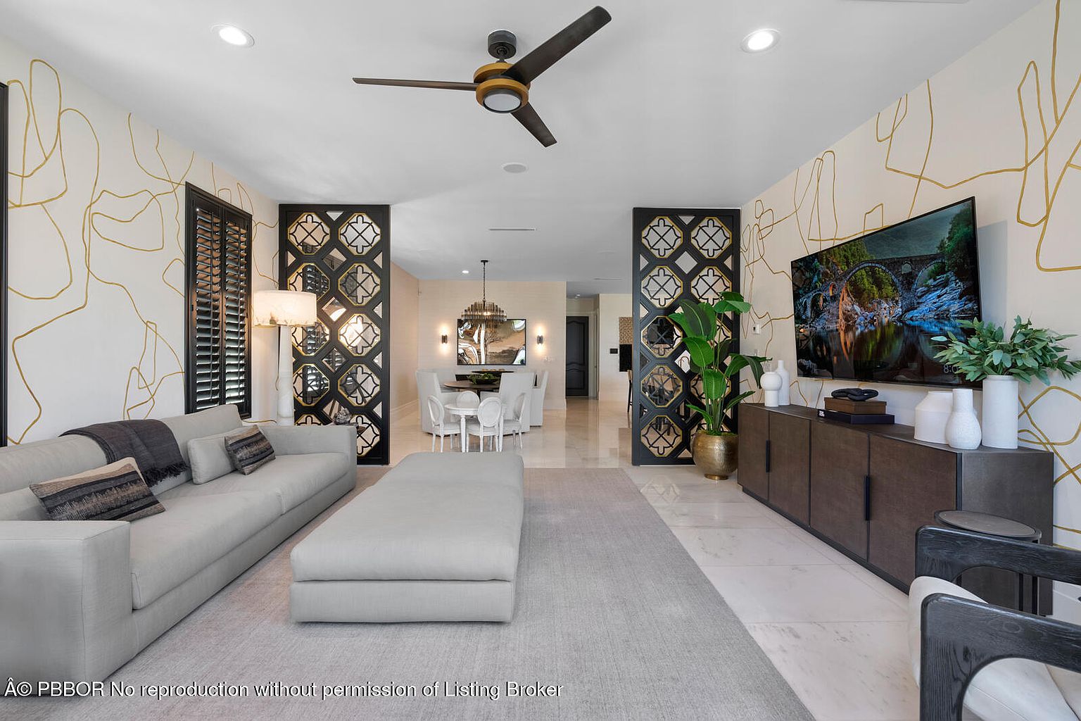 This is an interior shot of a modern living room featuring a neutral color palette with gold accents. The room is furnished with a large sectional sofa, an ottoman, and a media console with a large television. Decorative screens flank the opening to the dining area, and the walls are adorned with a contemporary gold line design, creating a sophisticated and inviting atmosphere.