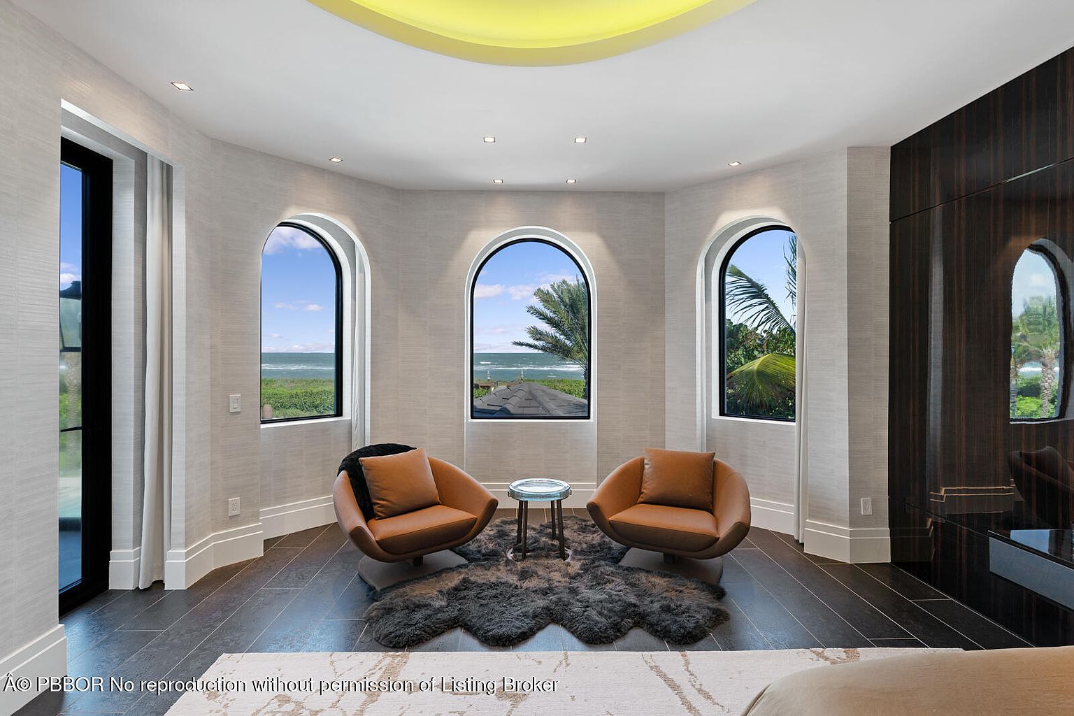 This interior shot showcases a stylish living room with a focus on natural light and modern design. The room features three arched windows offering ocean views, two contemporary brown leather chairs, and a unique fur rug. Dark wood flooring and a light-colored textured wall covering add to the room's sophisticated ambiance.