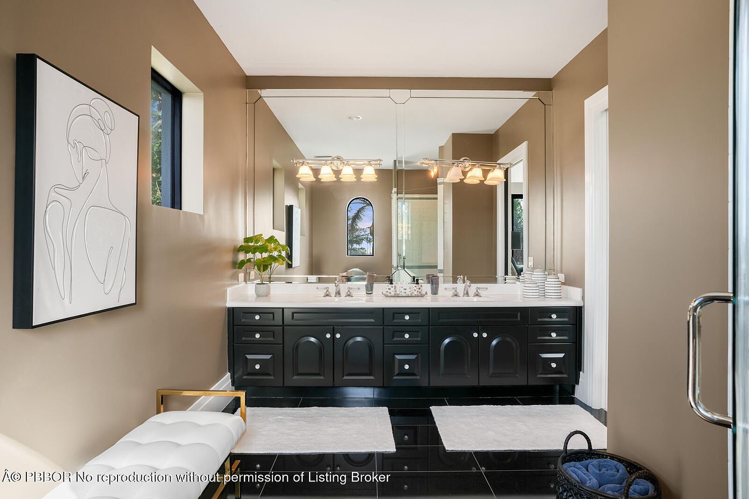 This is a primary bathroom featuring a double vanity with black cabinetry and a white countertop. A large mirror spans the length of the vanity, reflecting the lighting fixtures above. The floor is black tile, and there are white rugs in front of the vanity. A white bench is on the left side of the image and a basket of towels is on the right.