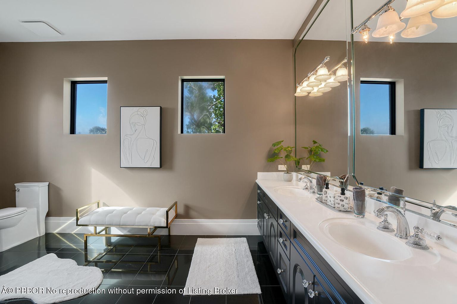 This is a primary bathroom featuring a double vanity with white countertops and dark blue cabinetry. The walls are painted in a neutral tone, and there are two small windows providing natural light. A large mirror reflects the vanity and the opposite wall, which includes a toilet, a bench, and artwork. The floor is black tile with white rugs.