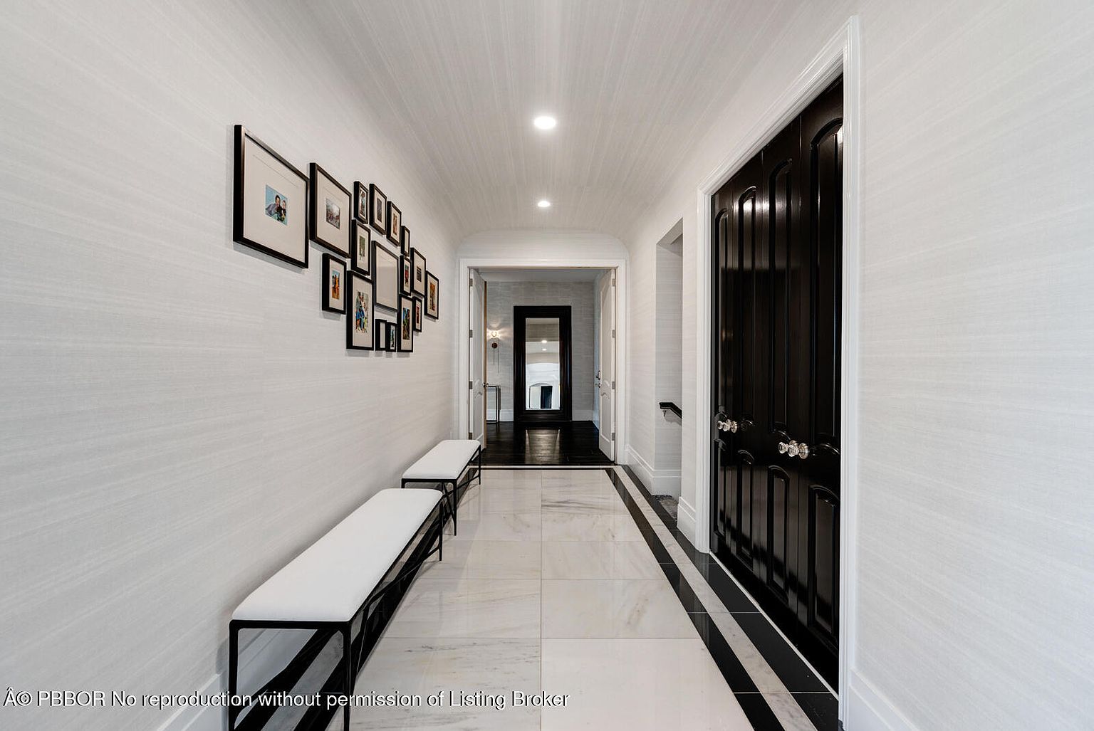 This is an interior shot of a hallway featuring a black and white color scheme. The hallway has white walls adorned with framed pictures and two benches with white cushions. The floor is tiled in white with black accents, and a black door is visible on the right. The perspective is a straight-on view, emphasizing the length of the hallway and the symmetry of the design.