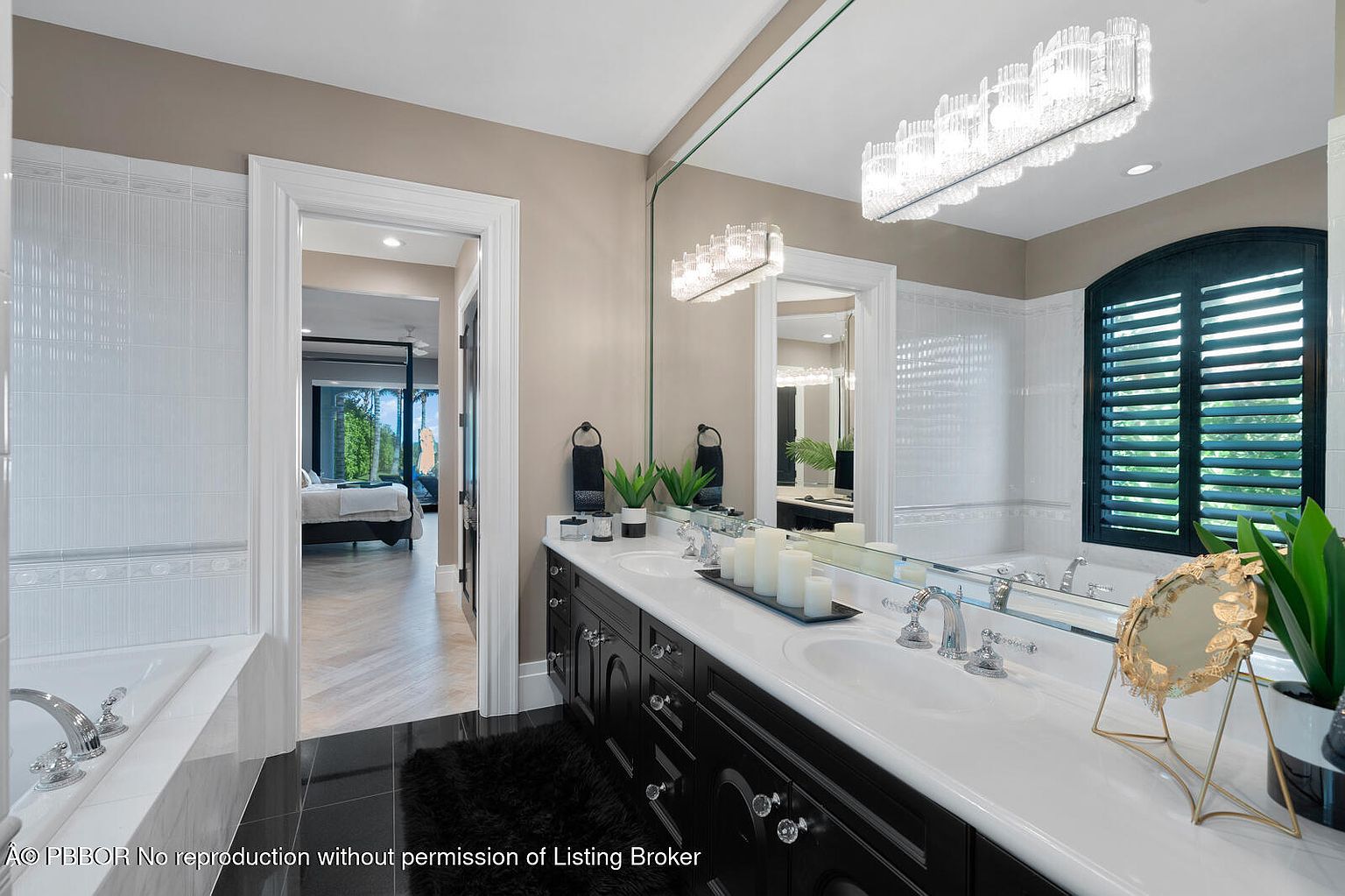This is a primary bathroom featuring a double vanity with a black cabinet base and white countertop, illuminated by modern crystal light fixtures. A large mirror spans the vanity, reflecting the space and enhancing the sense of openness. The bathroom also includes a soaking tub and a view into the primary bedroom, creating a luxurious and connected suite.