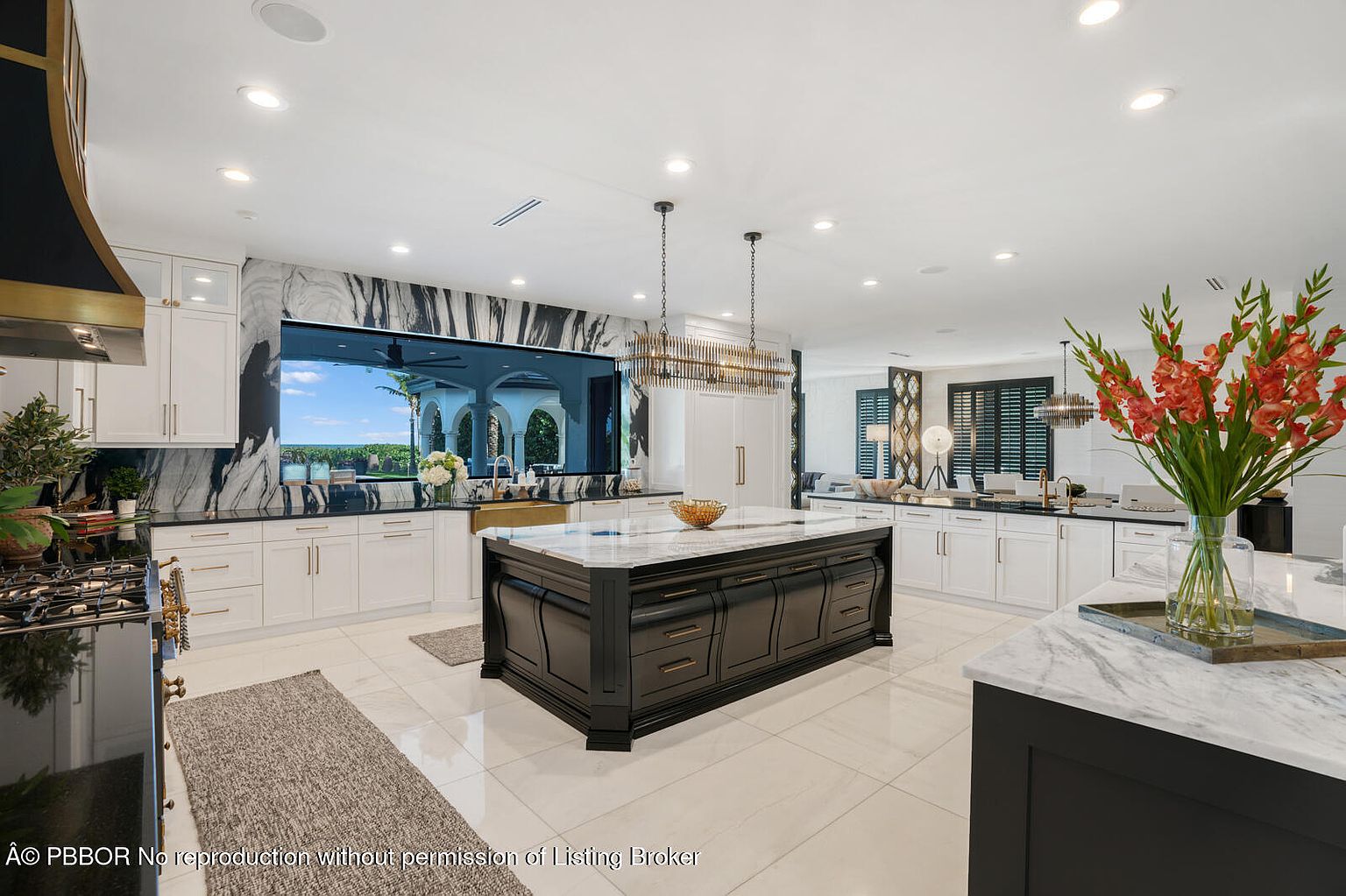 This is a bright and spacious kitchen featuring a large center island with a marble countertop and dark wood cabinetry. The kitchen is equipped with white cabinets, black countertops, and a large window offering a view of the exterior. The flooring is white tile, and the overall design is modern and luxurious.