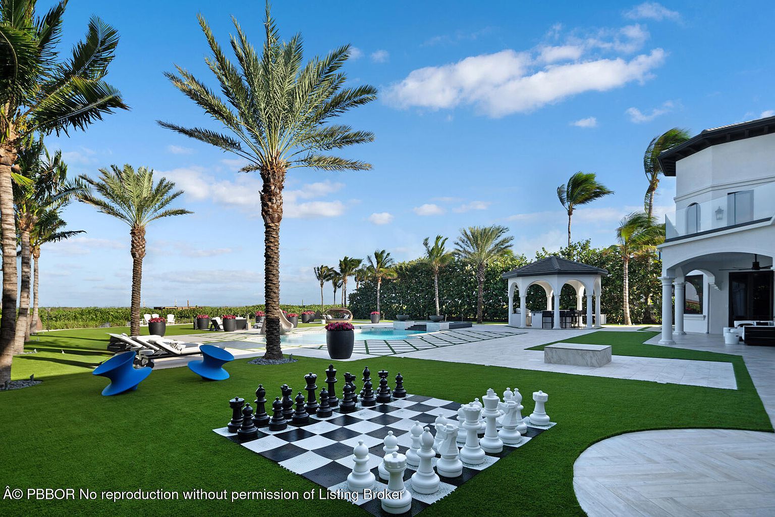 This exterior shot showcases a meticulously landscaped backyard featuring a large chessboard, palm trees, and a swimming pool. The property includes a gazebo and a glimpse of the home's architecture, suggesting a luxurious and well-maintained outdoor living space. The scene is set under a bright blue sky with scattered clouds, enhancing the overall appeal.