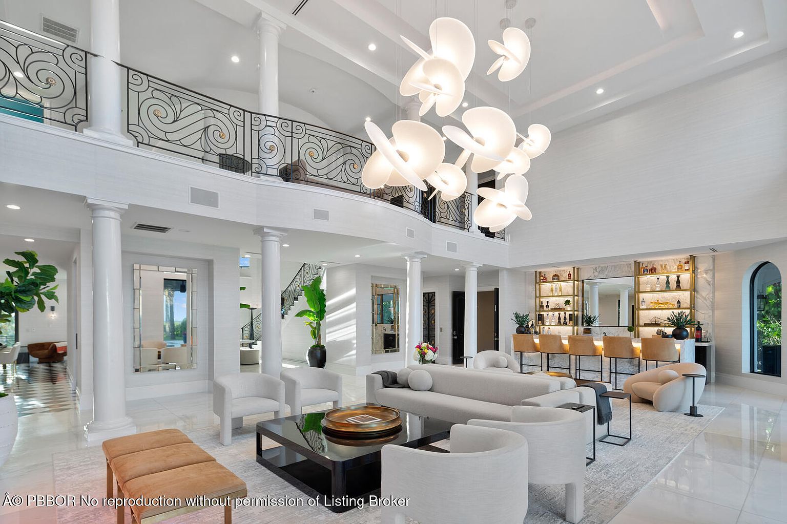 This is a grand living room featuring soaring ceilings and a large, modern chandelier. The space is decorated in neutral tones with white armchairs, a gray sofa, and a black coffee table. A bar area is visible in the background, and a balcony overlooks the living room, creating a luxurious and open atmosphere.