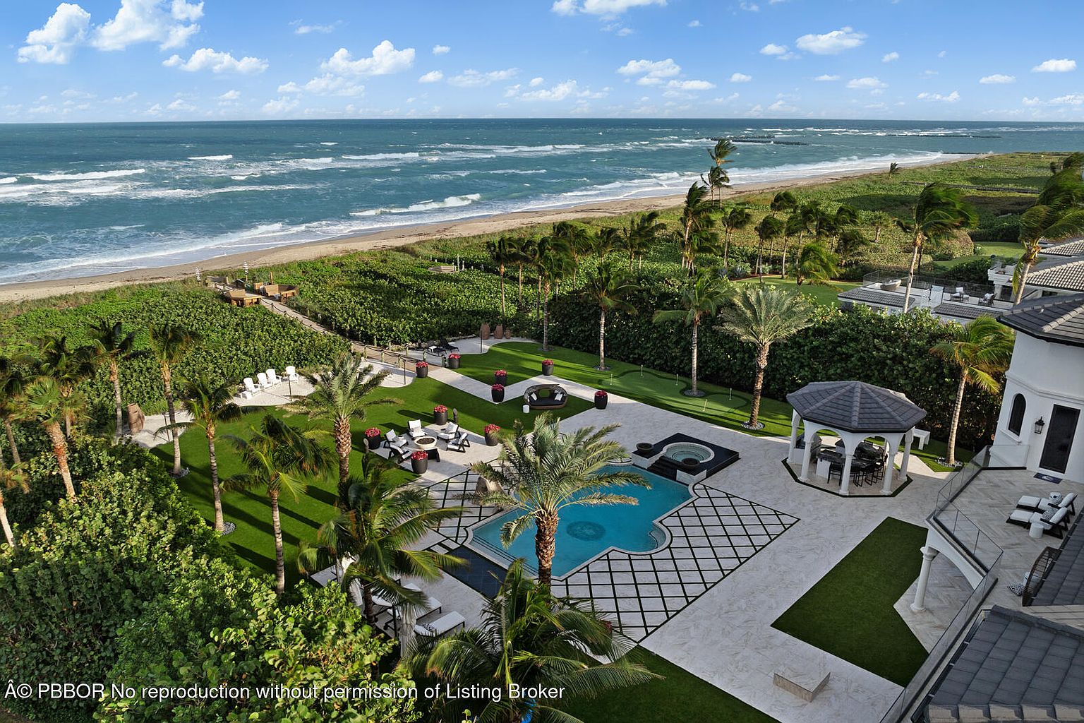 This aerial view showcases a luxurious beachfront property with a stunning pool and spa area, surrounded by meticulously manicured lawns and lush tropical landscaping. The property features a gazebo, a putting green, and direct access to a pristine beach, offering a serene and private oasis. The ocean provides a picturesque backdrop, enhancing the overall appeal of this high-end estate.