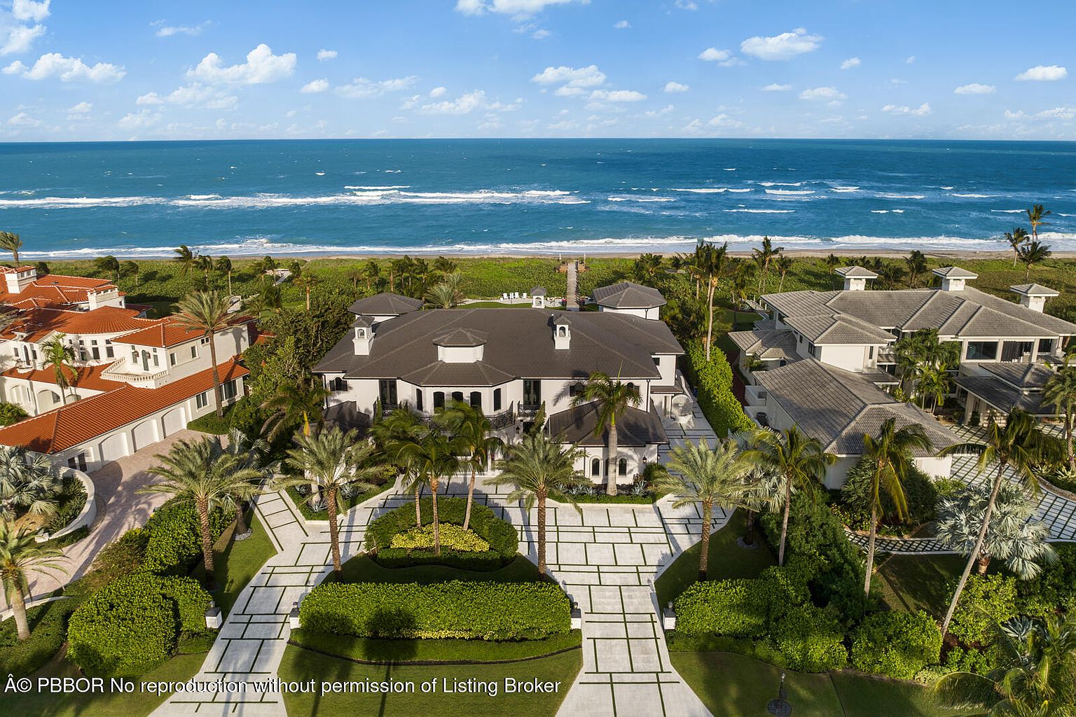 This aerial shot showcases a luxurious beachfront property with meticulously landscaped grounds. The grand residence features a dark roof, white exterior, and a symmetrical design, complemented by palm trees and manicured lawns. The property offers direct access to a pristine beach and ocean, creating an exclusive and serene living environment.