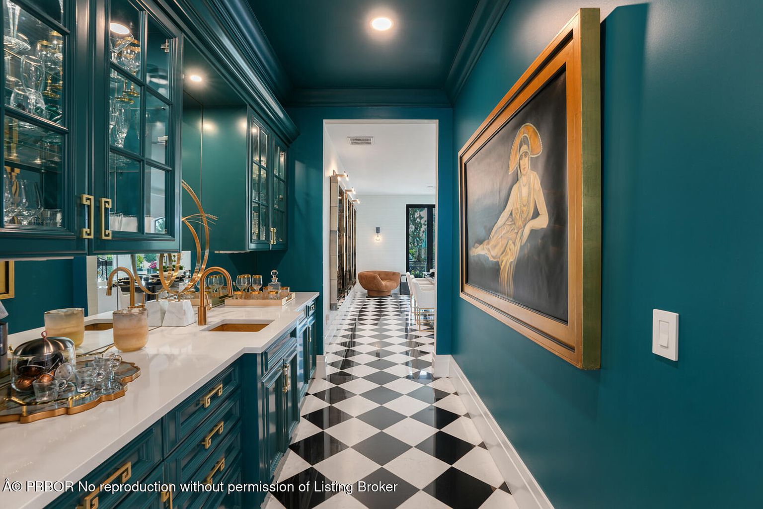 This interior shot showcases a stylish hallway featuring a wet bar with teal cabinetry, gold hardware, and white countertops. The hallway is adorned with a large framed painting and boasts a striking black and white checkered floor, leading to a glimpse of a living space. The overall aesthetic is luxurious and sophisticated.