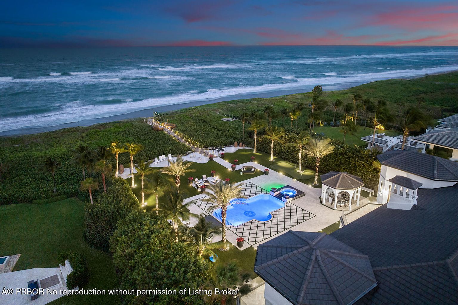 This aerial view showcases a luxurious beachfront property at dusk, featuring a meticulously landscaped backyard with a pool, spa, and gazebo. The property boasts direct access to the beach, with a lit pathway leading to the ocean. The overall impression is one of opulence and tranquility, highlighting the property's prime location and high-end amenities.