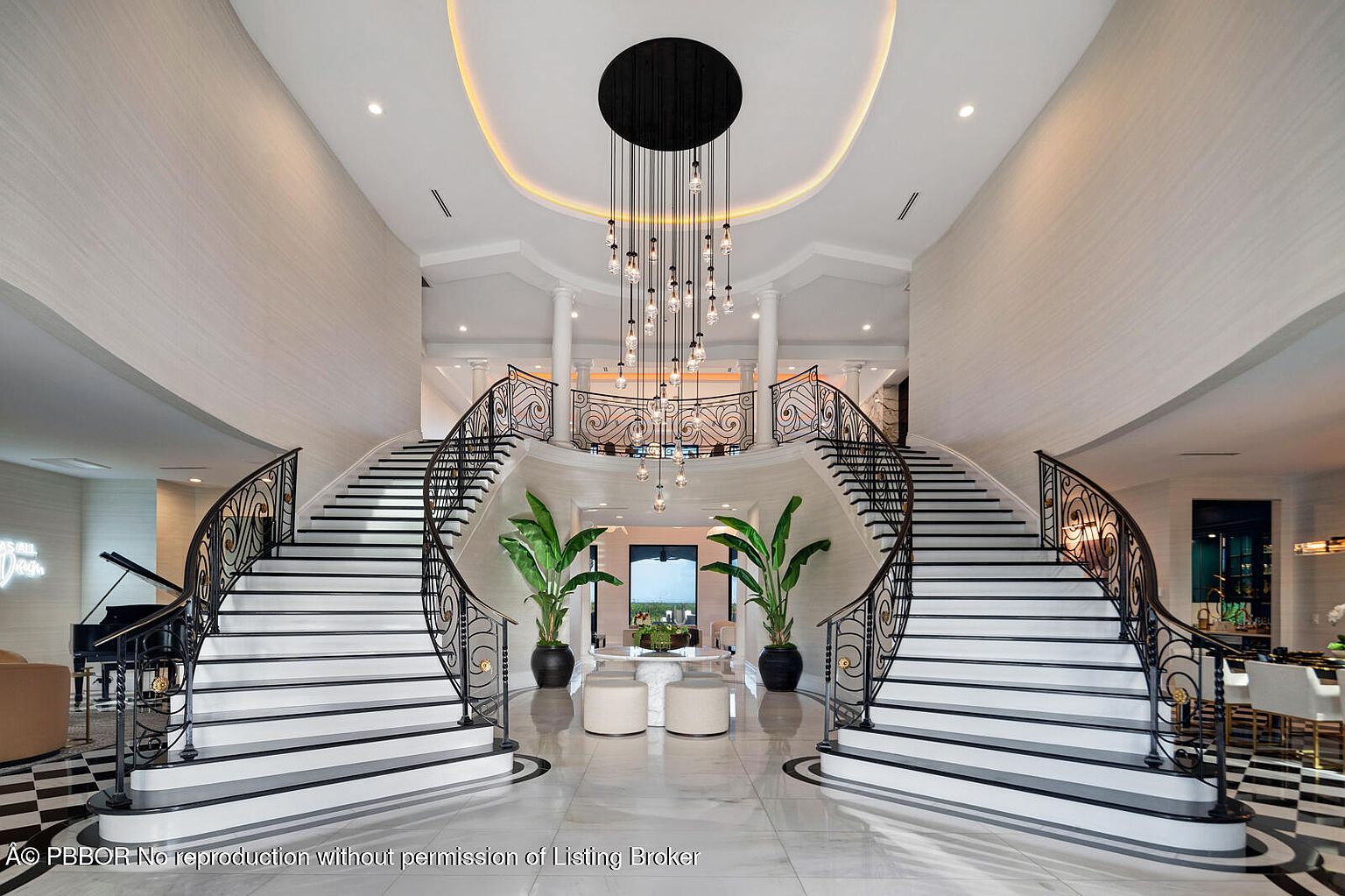 This grand foyer features dual curved staircases with ornate black railings and white treads, leading to an upper level. A striking chandelier hangs from the high ceiling, illuminating the marble flooring and potted plants that add a touch of nature. The space exudes luxury and elegance, creating a memorable first impression.