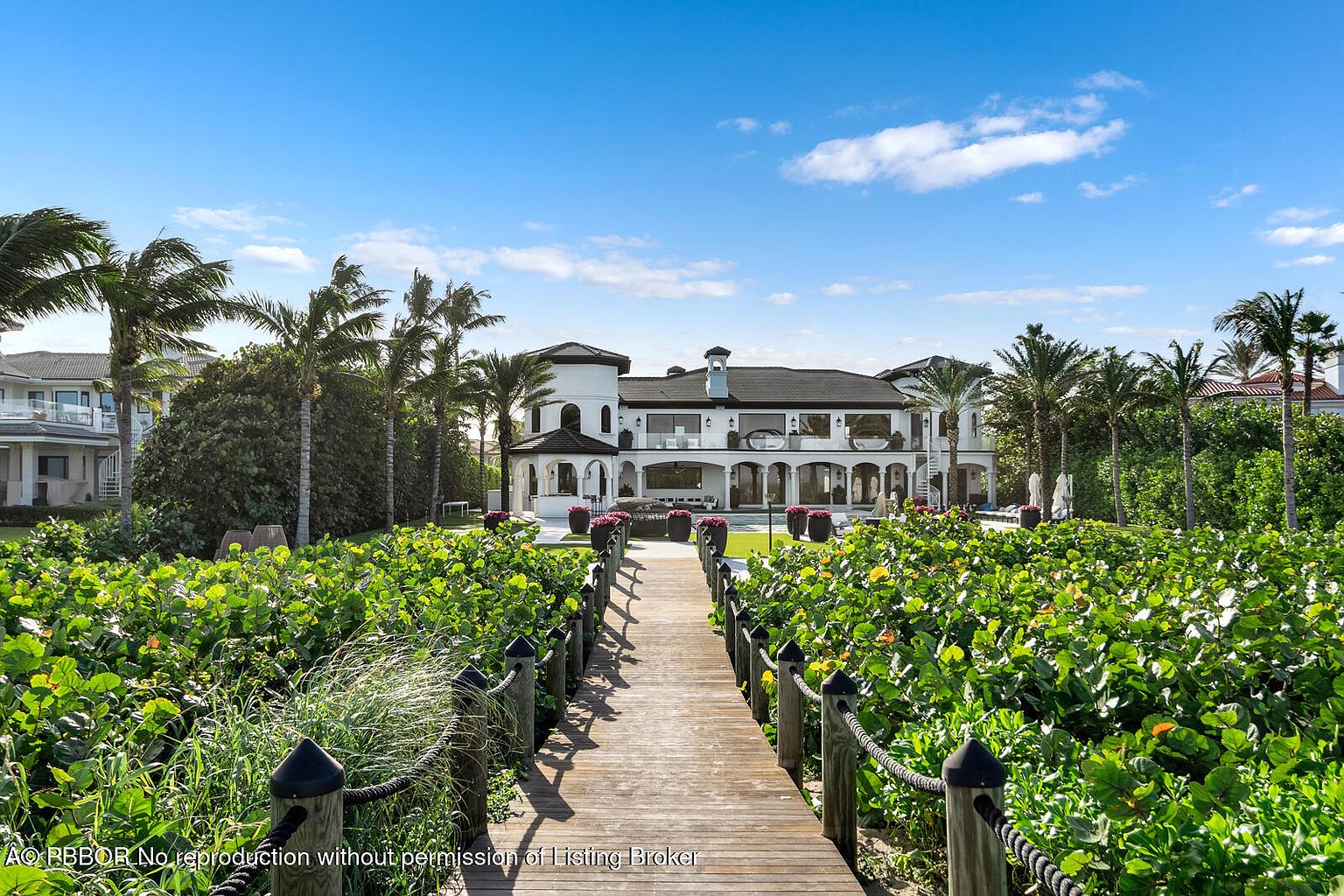 This image showcases a luxurious waterfront property, emphasizing its meticulously landscaped yard and garden. A wooden boardwalk, lined with rope railings, leads towards the grand white mansion, framed by lush greenery and palm trees. The scene evokes a sense of tranquility and exclusivity, highlighting the property's connection to nature and upscale living.