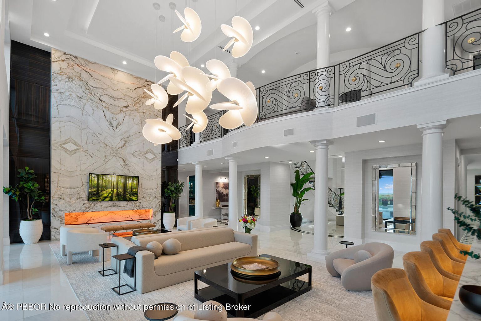 This is a luxurious living room featuring a high ceiling with a unique chandelier, a marble accent wall with a large television, and elegant furnishings. The space is bright and airy, with white columns and a balcony overlooking the area, creating a grand and sophisticated atmosphere. The color palette is neutral with pops of color from the artwork and plants.