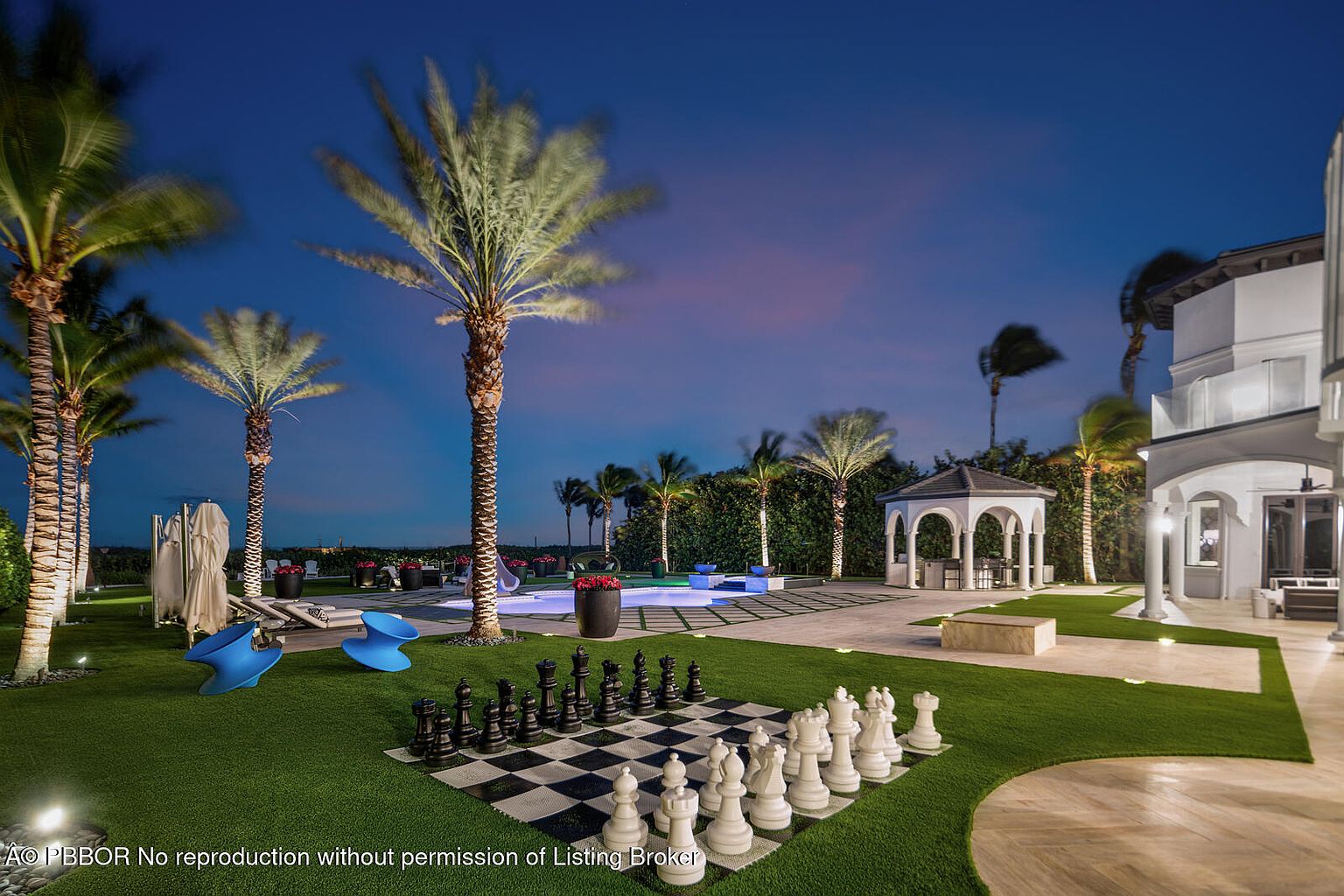 This exterior shot showcases a beautifully landscaped backyard at dusk, featuring a large chess set on the lawn, palm trees, and a pool area with lounge chairs. A gazebo and part of the house are visible in the background, creating an inviting and luxurious outdoor living space. The perspective is from a low angle, emphasizing the depth and grandeur of the yard.