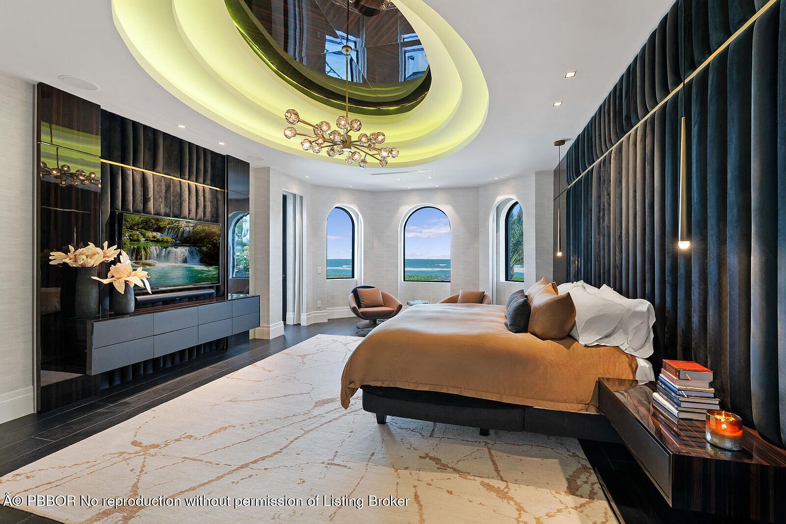 This is a luxurious primary bedroom featuring a unique circular ceiling design with integrated lighting and a modern chandelier. The room boasts a large bed with a plush headboard, complemented by stylish nightstands and elegant decor. Arched windows offer a view of the ocean, enhancing the room's serene and upscale ambiance.