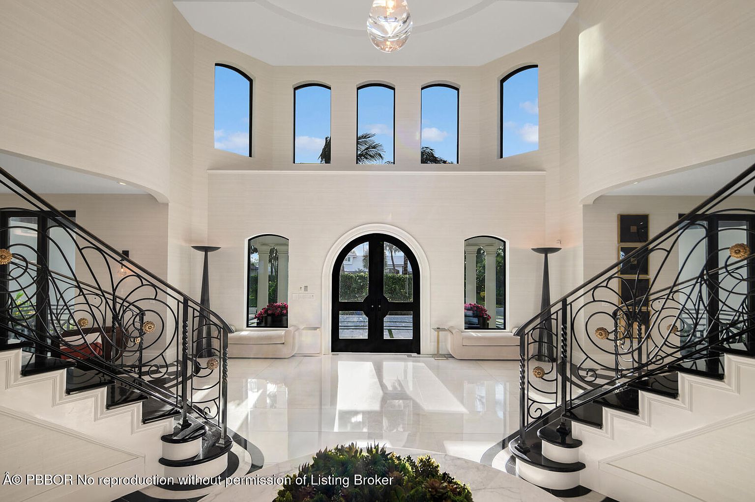 This grand foyer features dual staircases with ornate black iron railings and white marble steps, creating a symmetrical and elegant entrance. The high ceilings are accentuated by arched windows and a central chandelier, while the marble flooring adds a luxurious touch. The overall impression is one of spaciousness and sophisticated design.