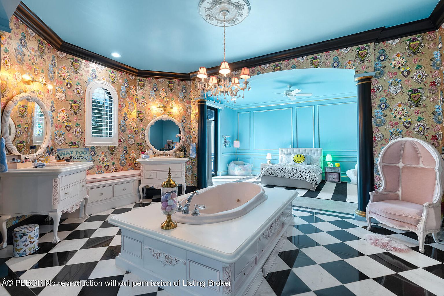 This is a whimsical and luxurious primary bathroom featuring a black and white checkered floor, ornate white vanities with round mirrors, and a large soaking tub in the center of the room. The walls are covered in patterned wallpaper, and the adjacent bedroom area is painted in a bright blue, creating a playful and unique space. The room is well-lit by a chandelier and wall sconces.