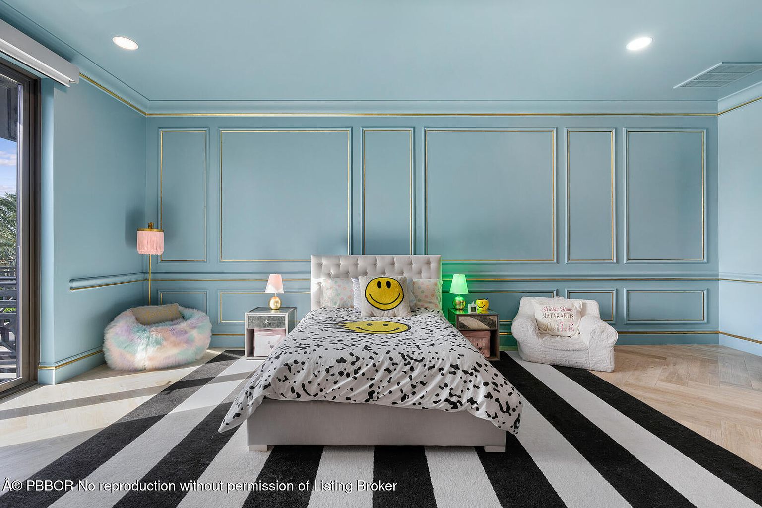 This is a stylish bedroom featuring a light blue paneled wall with gold trim, creating a sophisticated and playful atmosphere. The room is furnished with a bed adorned with smiley face pillows and a patterned duvet, flanked by mirrored nightstands and lamps. A black and white striped rug adds a bold contrast to the light-toned walls and flooring, enhancing the room's modern aesthetic.