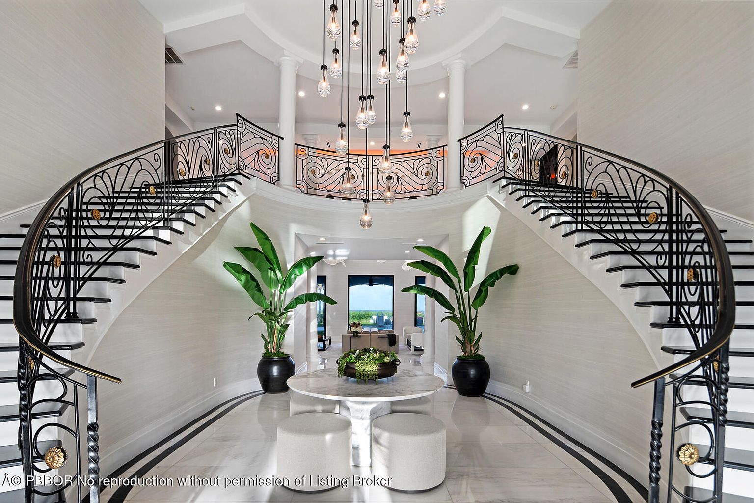 This grand foyer features dual curved staircases with ornate black metal railings and gold accents, flanking a central area with a round marble table and potted plants. A striking chandelier hangs from the high, domed ceiling, and the marble flooring adds to the luxurious feel. The space is bright and airy, creating a dramatic entrance.