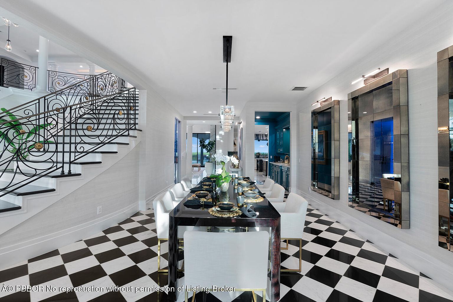 This interior shot showcases a sophisticated dining room with a striking black and white checkered floor. A long, dark wood dining table with gold accents is set for a formal meal, surrounded by white chairs. The room is well-lit by a modern chandelier and features decorative mirrors on the wall, adding depth and elegance to the space, while a staircase is visible on the left.