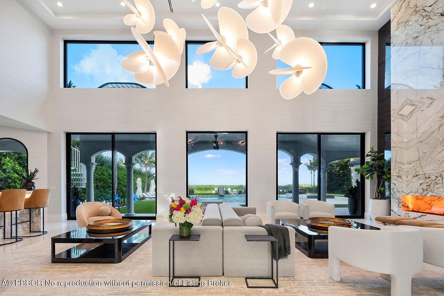 This is a luxurious living room featuring high ceilings, large windows with views of the outdoors, and modern lighting fixtures. The room is furnished with a neutral-toned sectional sofa, armchairs, and sleek coffee tables. A marble fireplace adds a touch of elegance, creating a sophisticated and inviting atmosphere.