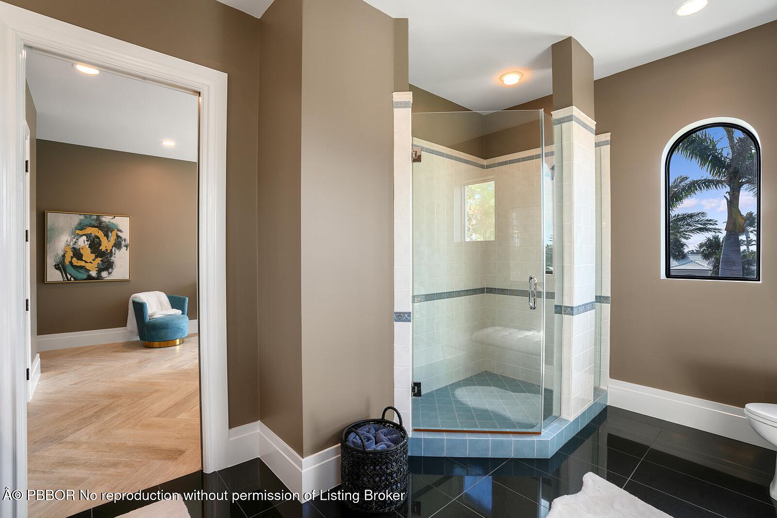 This is a primary bathroom featuring a glass-enclosed shower with light-colored tile and a blue accent border. The bathroom has dark tile flooring, and the walls are painted in a neutral tone. An arched window provides natural light, and a glimpse into an adjacent room with a chair and artwork is visible through a doorway.