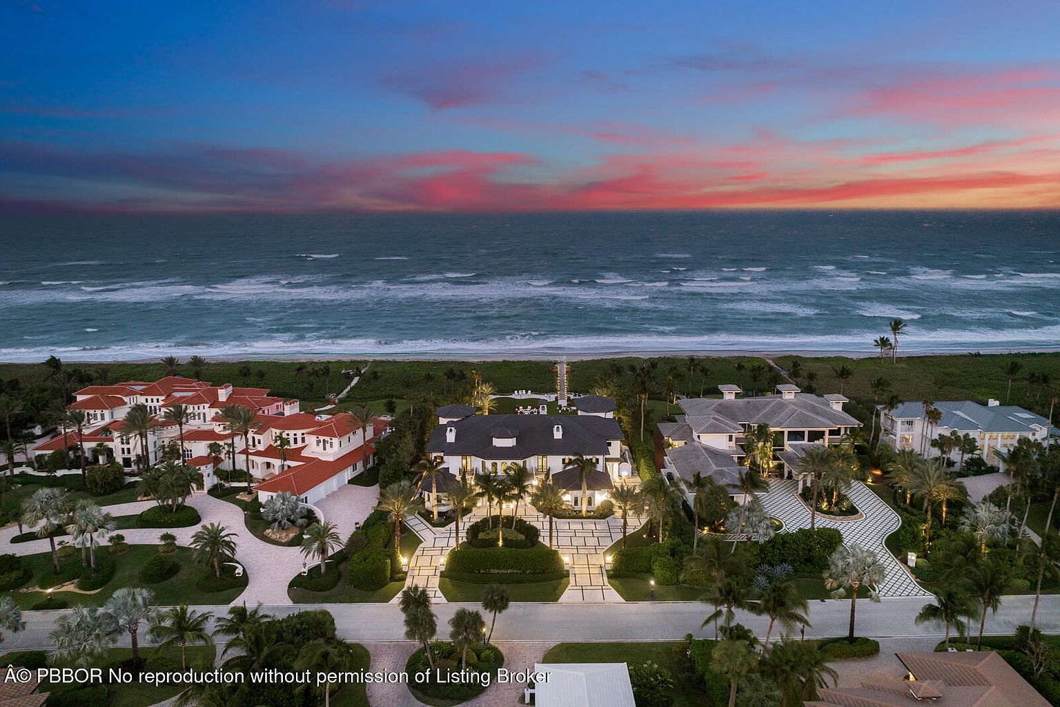 This aerial shot showcases a luxurious beachfront property at dusk, featuring multiple buildings with distinct architectural styles. The main residence boasts a dark roof and manicured landscaping, while neighboring structures exhibit red-tiled roofs and elegant designs. The ocean provides a stunning backdrop, enhancing the property's appeal and highlighting its prime location.