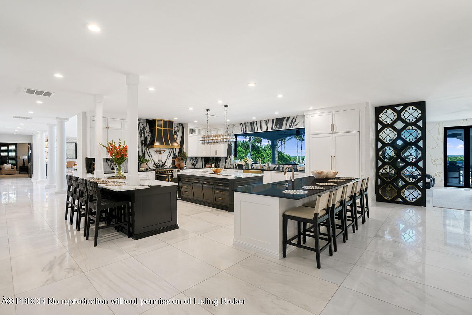 This is a spacious, modern kitchen featuring two large islands, one with seating. The kitchen boasts a black and white color scheme with marble accents and high-end appliances. Large windows provide natural light and views of the outdoors, enhancing the luxurious feel of the space.