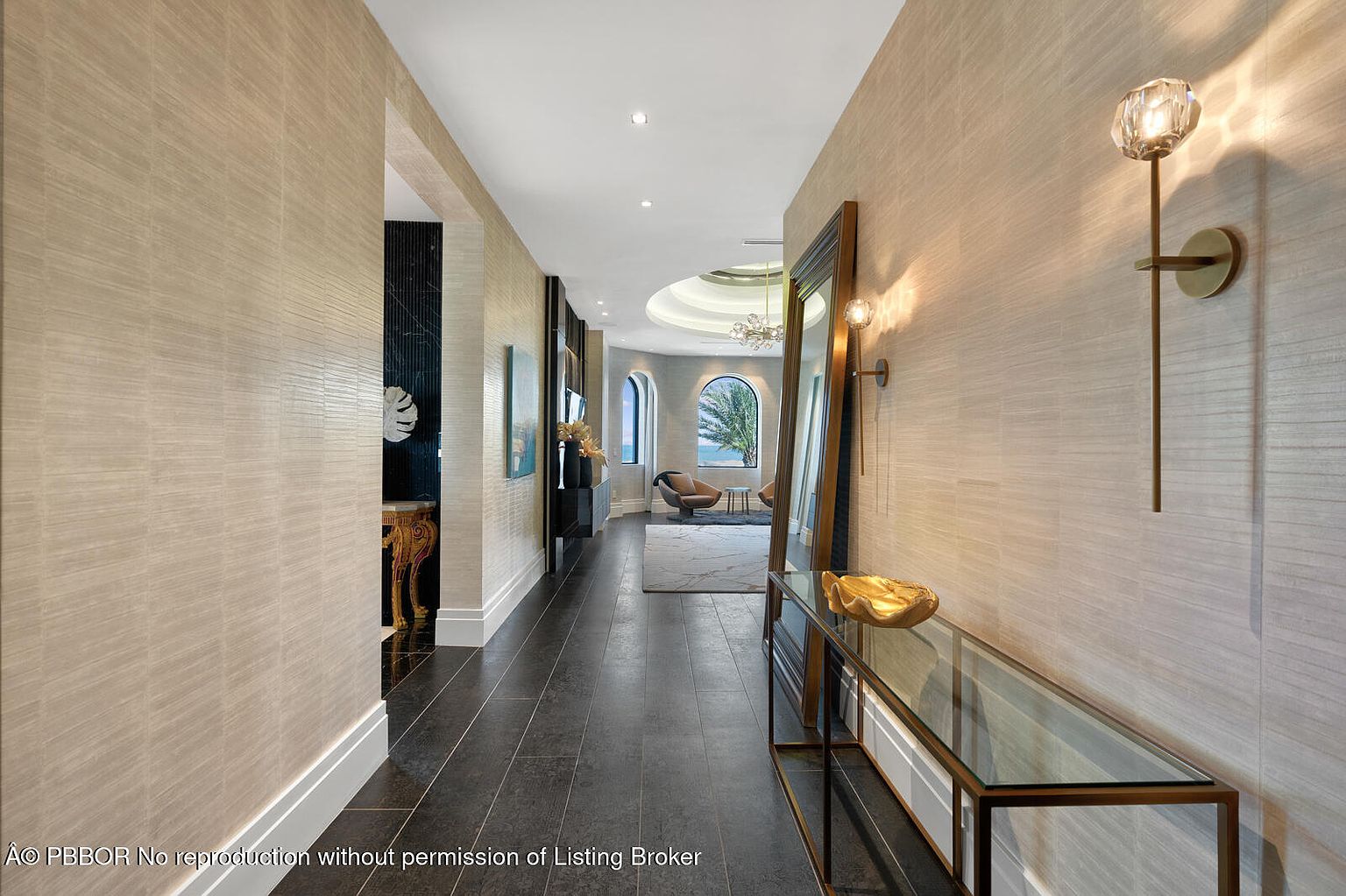 This is an interior shot of a luxurious hallway featuring dark wood flooring and textured beige wallpaper. A glass-topped console table with a gold accent piece sits against the wall, illuminated by a modern sconce. The hallway leads to a bright living area with arched windows, suggesting a high-end, sophisticated home.