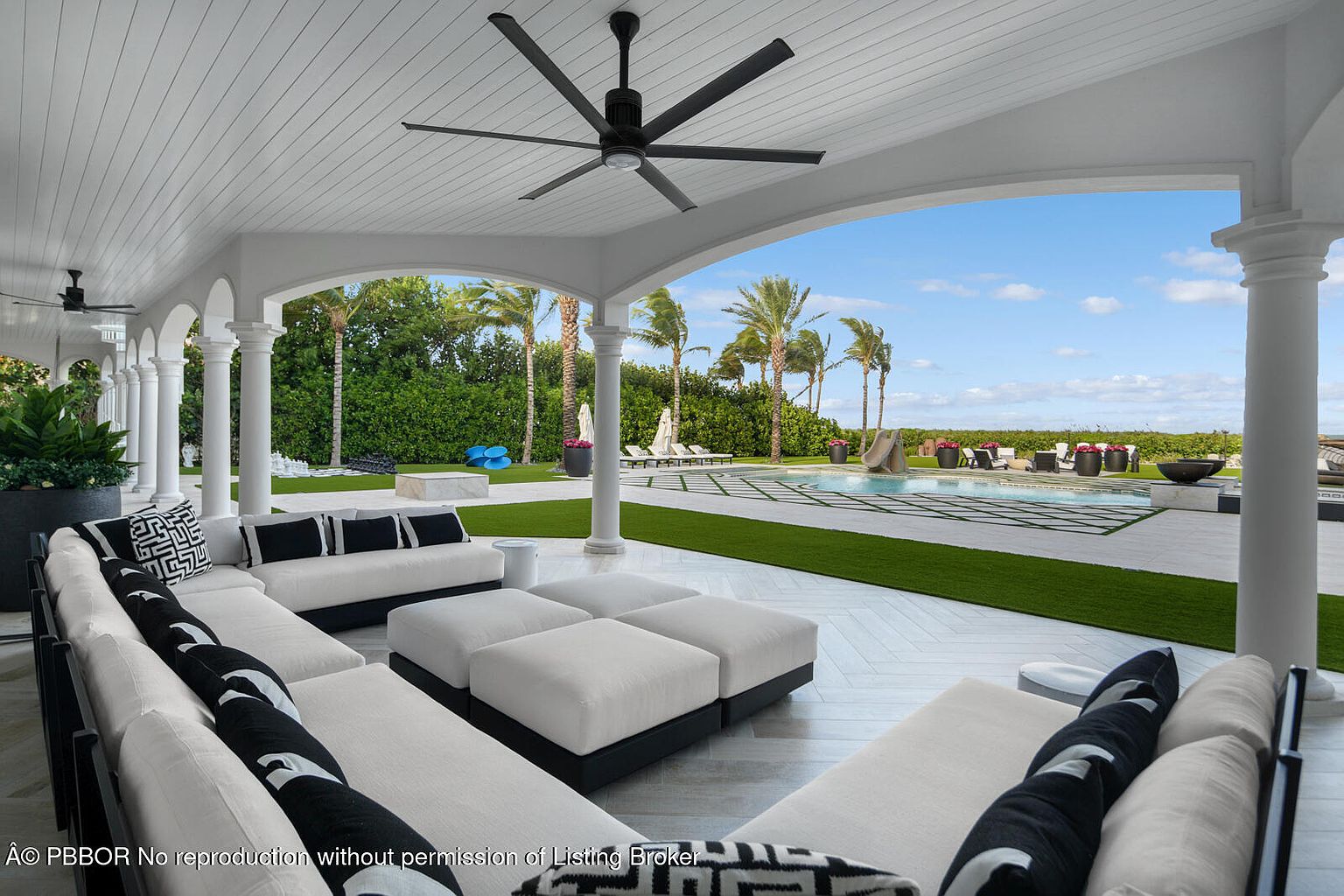 This image showcases a luxurious outdoor patio area with a covered seating space, featuring elegant white columns and a ceiling fan. The patio overlooks a beautifully landscaped backyard with a pool, palm trees, and manicured lawn. The furniture is modern and stylish, creating an inviting and upscale outdoor living space.