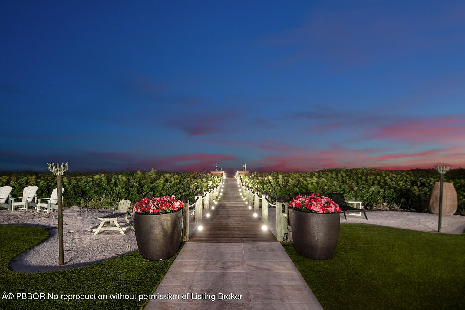 This image showcases a beautifully landscaped yard and garden area, featuring a pathway leading towards the beach. The pathway is lined with lush greenery, decorative planters filled with vibrant flowers, and elegant lighting fixtures. The overall impression is one of tranquility and sophistication, perfect for a luxury real estate listing.