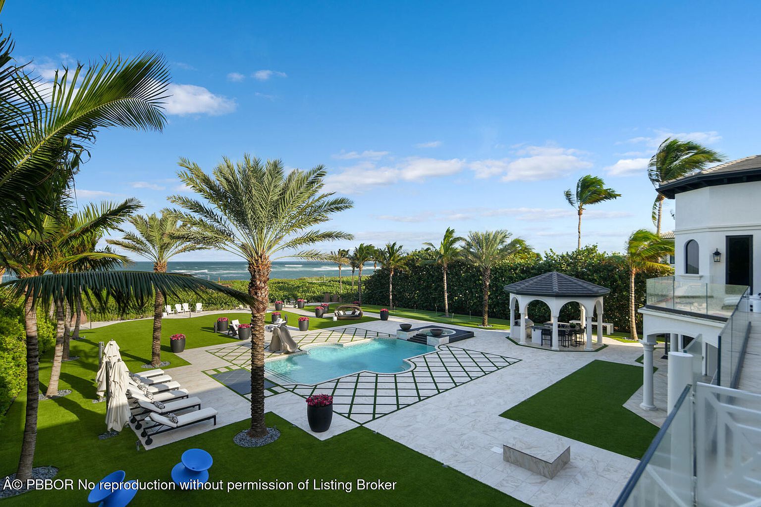 This is an exterior shot showcasing a luxurious pool and spa area. The pool is surrounded by a meticulously designed patio with geometric patterns and lush green grass. Palm trees line the perimeter, offering a tropical ambiance, and a gazebo provides a shaded seating area, enhancing the property's appeal for relaxation and entertainment.