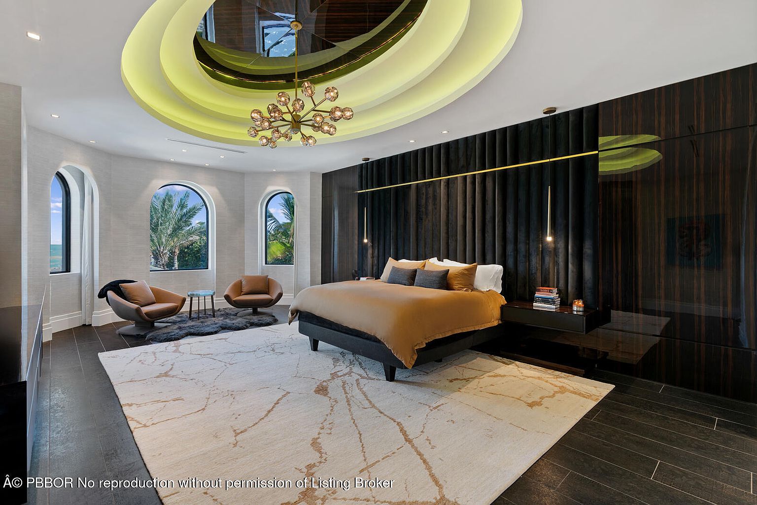 This is a luxurious primary bedroom featuring a unique circular ceiling with recessed lighting and a modern chandelier. The room includes a large bed with a dark headboard, a nightstand with books, and two armchairs near arched windows that offer natural light. A large area rug covers most of the dark wood flooring, adding warmth and texture to the space.