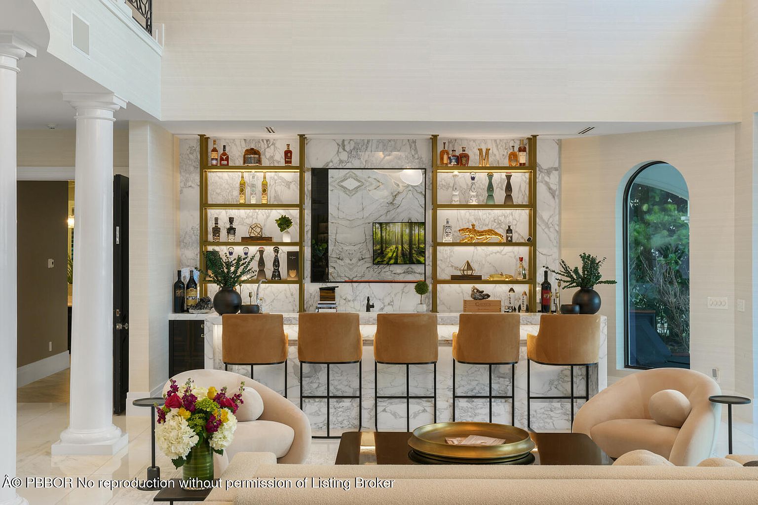 This interior shot showcases a stylish living room with a built-in bar area featuring marble accents and gold shelving. The bar is complemented by modern bar stools and a television, while comfortable seating arrangements create an inviting atmosphere. The room's design blends luxury with comfort, making it an ideal space for relaxation and entertainment.