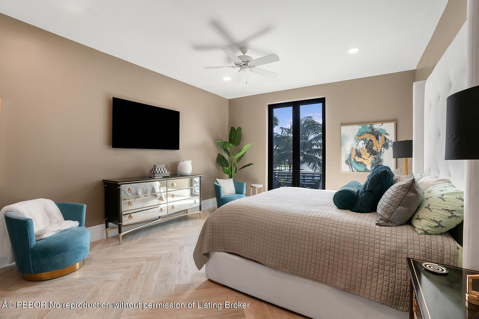 This is a primary bedroom featuring a large bed with a quilted coverlet and decorative pillows, a mirrored dresser with decorative items, and a blue accent chair. The room has a neutral color palette with pops of color from the furniture and artwork, and a herringbone wood floor adds texture. A black-framed window provides natural light and a view to the outside.