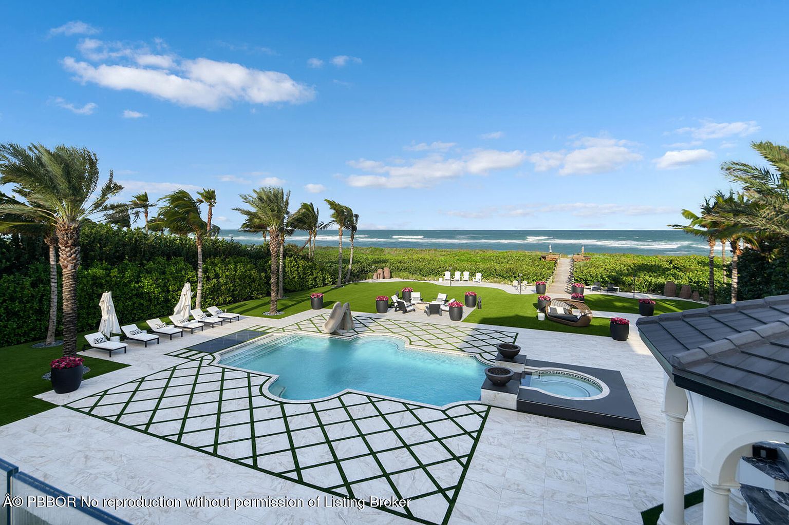 This image showcases a luxurious backyard with a stunning pool and spa area. The pool features a unique shape and is surrounded by a patterned tile patio with grass accents. Beyond the pool, there is a well-manicured lawn leading to the beach, complete with lounge chairs, a seating area, and lush landscaping, creating a private and serene oasis.