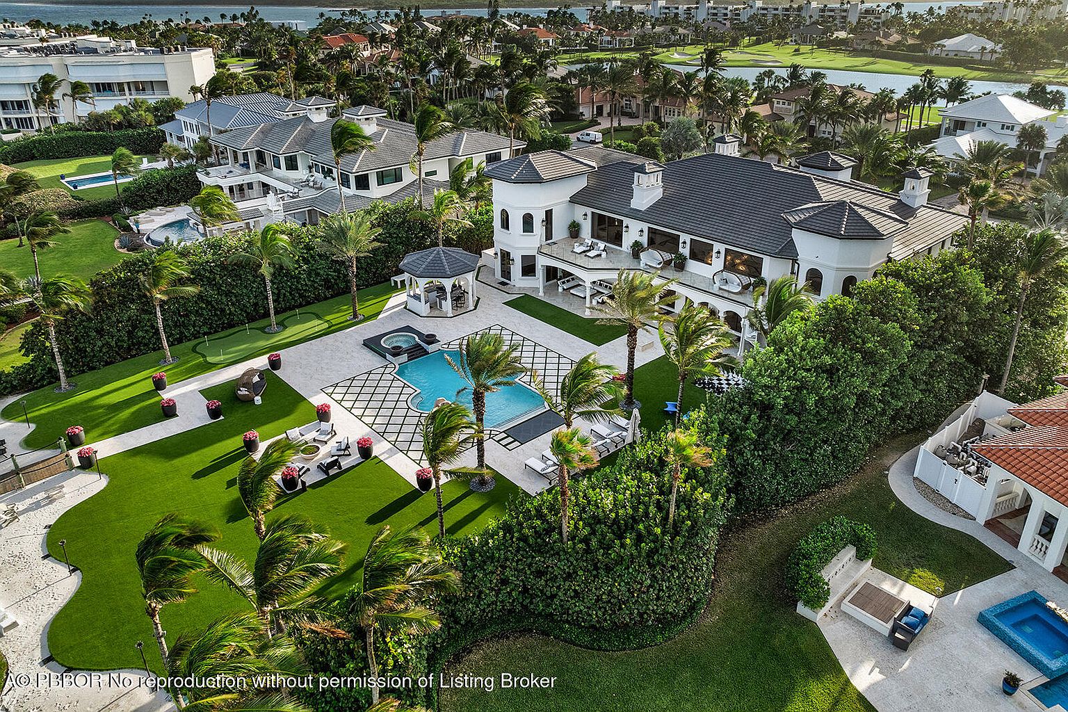 This aerial view showcases a luxurious estate with a large pool, meticulously landscaped grounds, and a grand residence featuring a turret and multiple outdoor living spaces. The property includes a gazebo, a putting green, and a well-manicured lawn, all set against a backdrop of palm trees and a waterfront view, creating an impression of opulence and privacy.