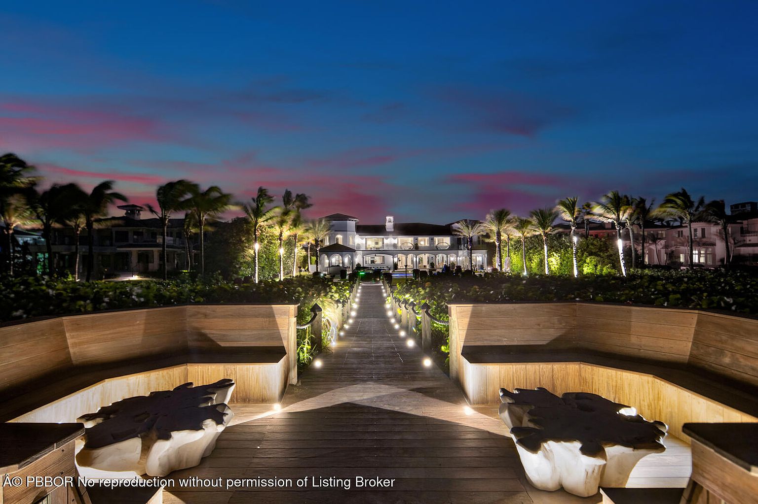 This image showcases the front exterior of a luxurious beachfront property at dusk. A wooden walkway, lined with lights and lush greenery, leads to the grand residence, which is framed by palm trees and a vibrant sky. The scene exudes elegance and tranquility, highlighting the property's prime location and upscale design.