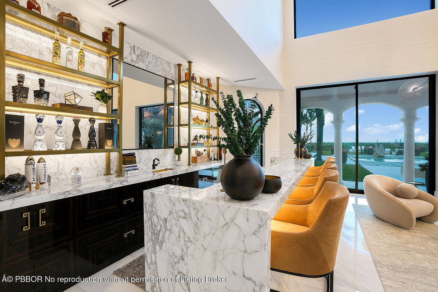 This interior shot showcases a stylish bar area within a luxurious living space. The bar features marble countertops, dark wood cabinetry with gold hardware, and illuminated shelving displaying various bottles. The room opens to an outdoor pool area through large sliding glass doors, creating a seamless indoor-outdoor living experience.