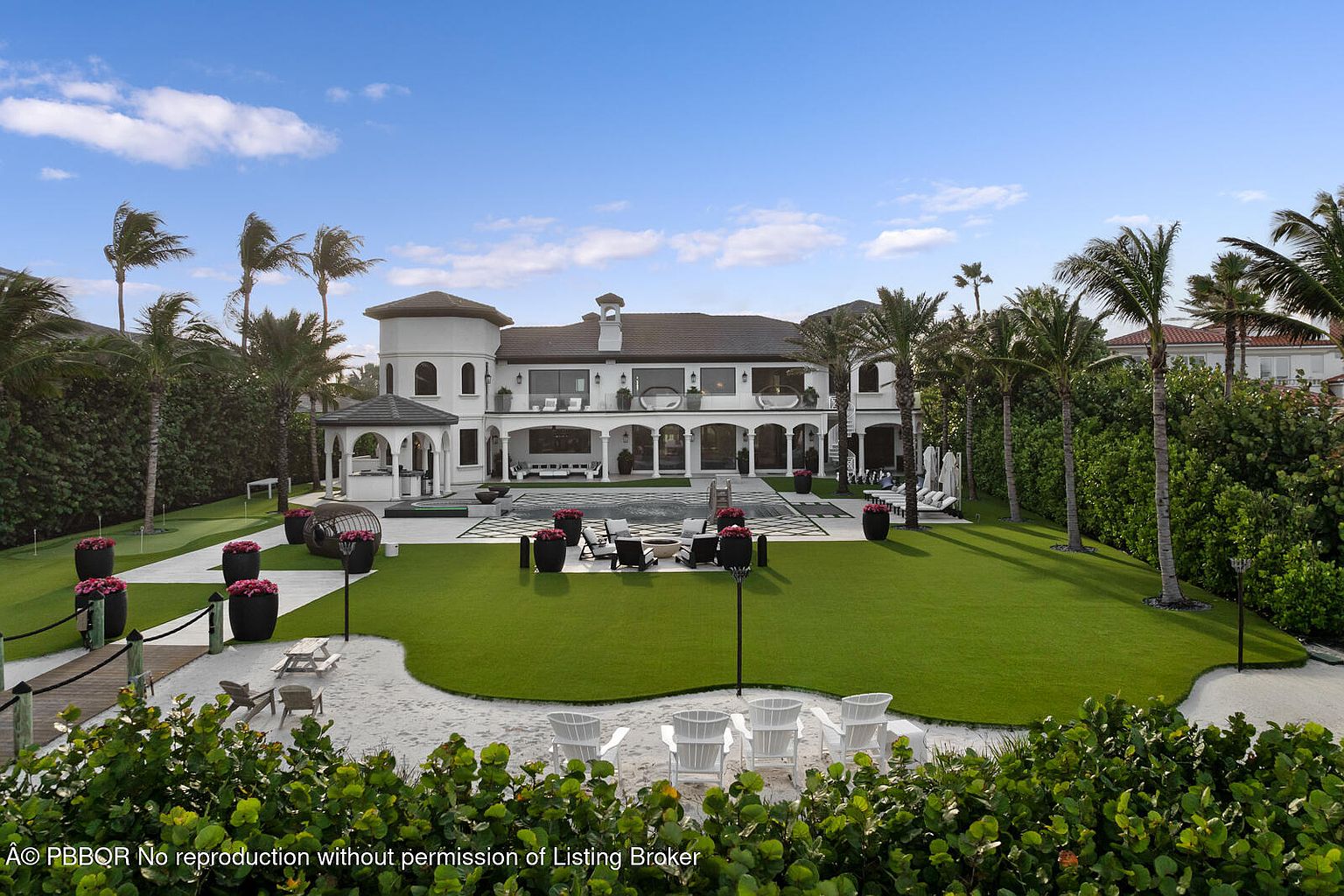 This image showcases the rear exterior of a luxurious estate, featuring a sprawling green lawn, a pristine pool area, and a sandy beach-like space with lounge chairs. The architecture is elegant with white columns, a tiled roof, and multiple balconies, complemented by lush landscaping including palm trees and manicured bushes. The overall impression is one of opulence and resort-style living.
