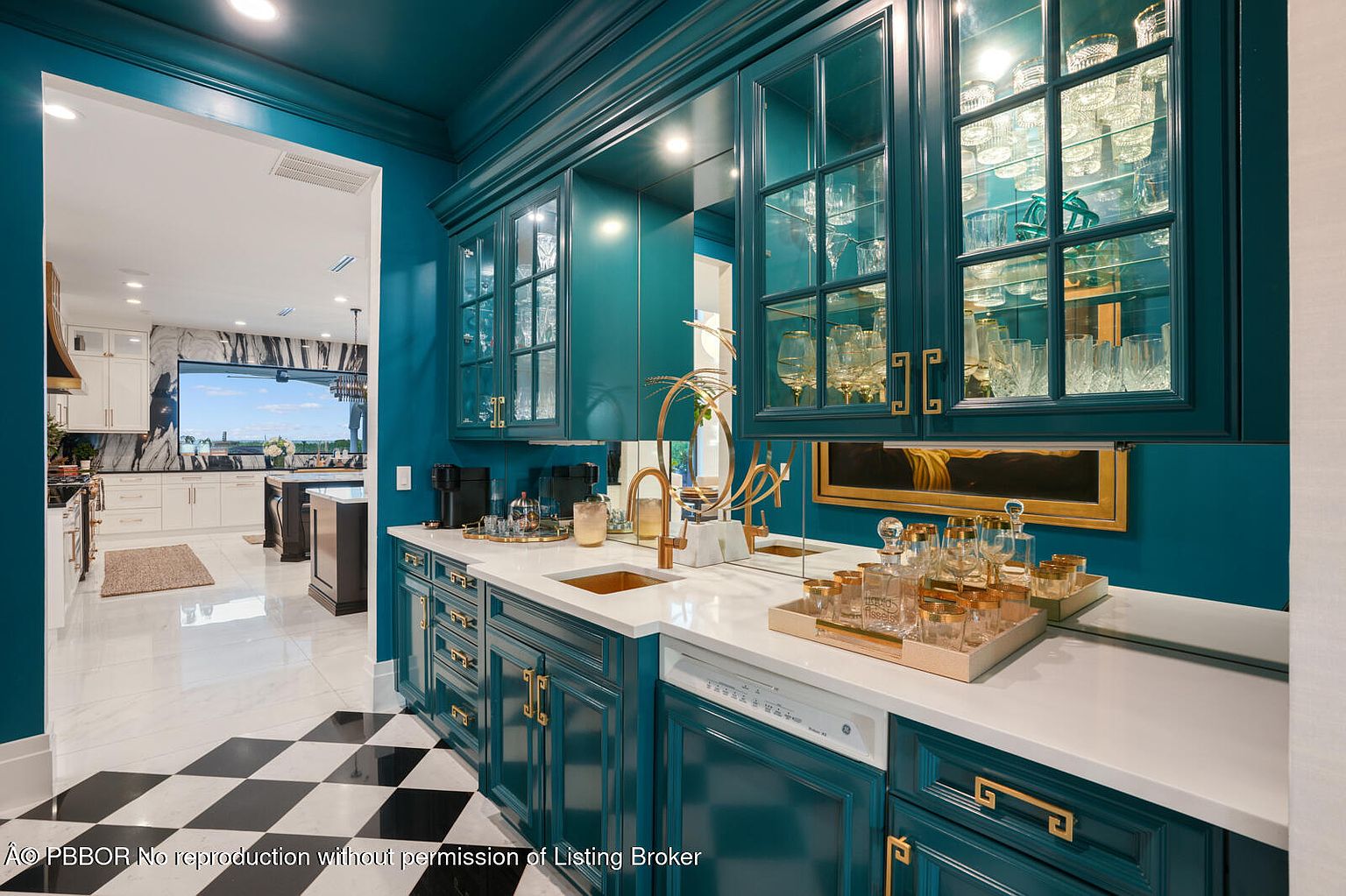 This image showcases a stylish kitchen area with custom teal cabinetry, gold hardware, and glass-front display cabinets filled with glassware. A white countertop complements the teal cabinets, and a copper sink adds a touch of luxury. The black and white checkered flooring adds a classic touch to the space.