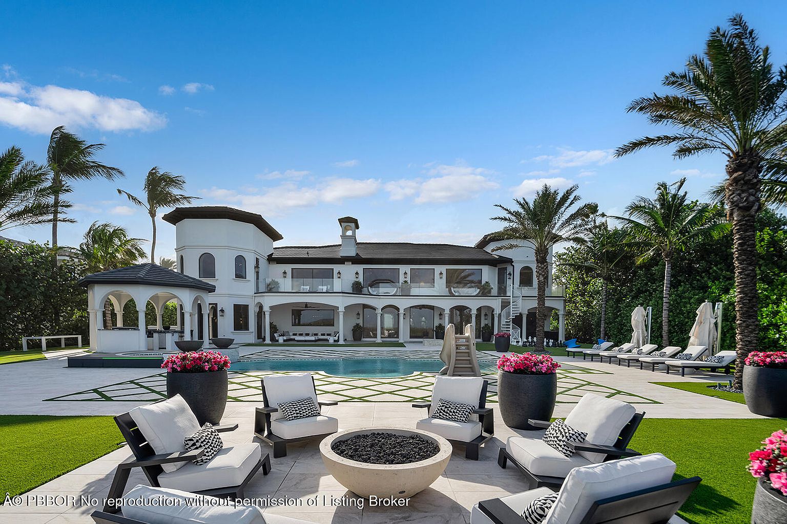 This image showcases the rear exterior of a luxurious home, featuring a pristine pool, elegant patio furniture arranged around a fire pit, and meticulously manicured landscaping. The architectural style is sophisticated, with white walls, dark roofing, and arched openings that create a seamless transition between indoor and outdoor living spaces. Palm trees and lush greenery add to the property's tropical ambiance, enhancing its appeal as a high-end real estate offering.