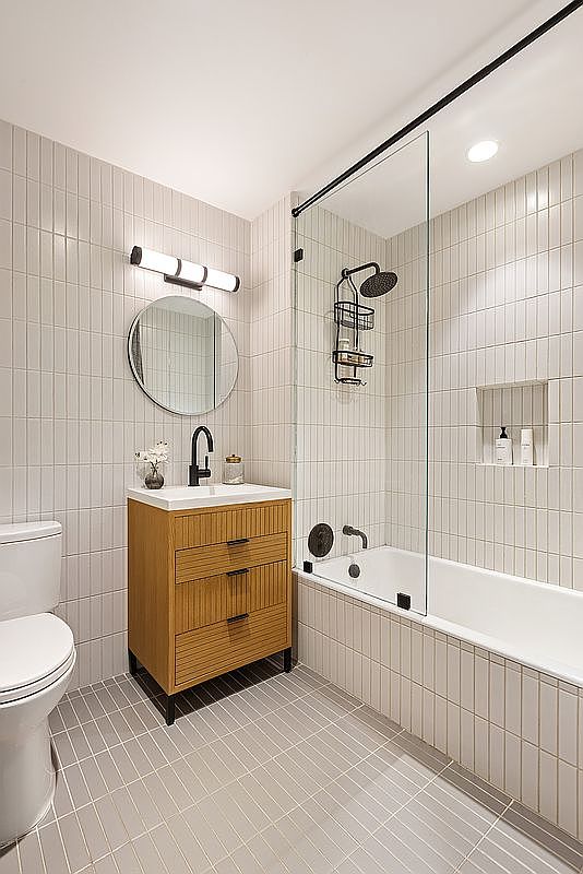 This is a well-lit bathroom featuring a modern design with vertical white tiles on the walls and floor. A wooden vanity with black hardware and a round mirror are prominent, along with a toilet and a bathtub enclosed by a glass shower screen. The overall impression is clean, stylish, and functional.