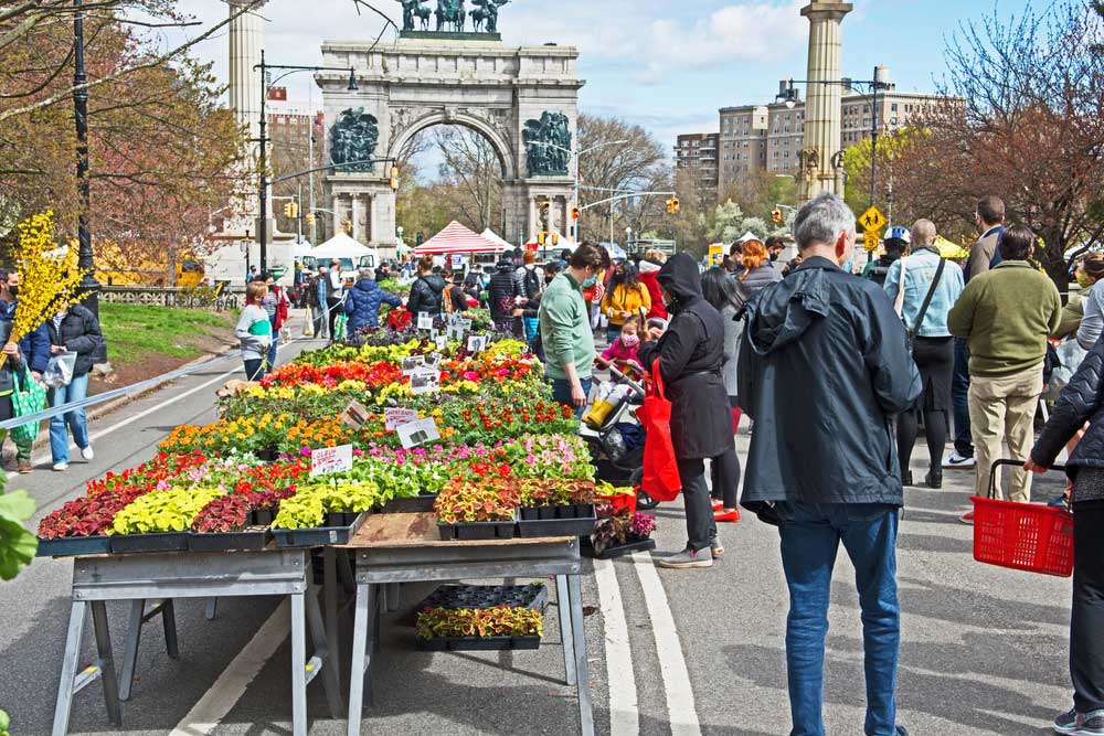 This image showcases a bustling outdoor market held in a park. Colorful flower displays are prominent along the street, attracting numerous visitors. Arched architectural elements and cityscape features are visible in the background, contributing to the vibrant and lively atmosphere and hinting at the location's urban setting.