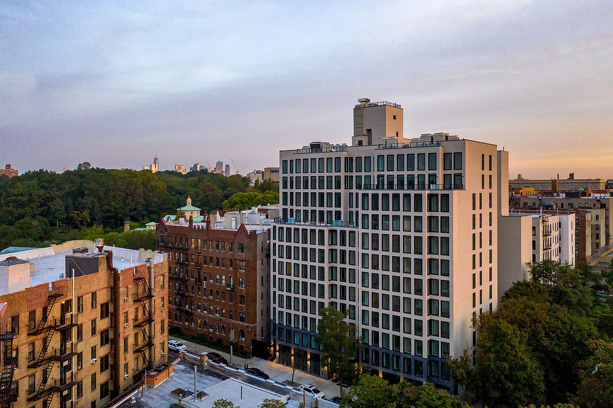 The image provides an aerial view of several buildings, including a modern white apartment building with numerous windows and a red-brick building with fire escapes. The architectural style is a mix of old and new urban design. In the distance, a city skyline is visible beyond a green, tree-filled area.