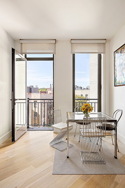 This is an interior shot of a dining room featuring a glass-top table with metal frame and four metal chairs. The room has two large windows with light-filtering shades, one of which leads to a balcony. A light gray rug sits beneath the table, and a piece of artwork hangs on the wall, creating a bright and modern dining space.