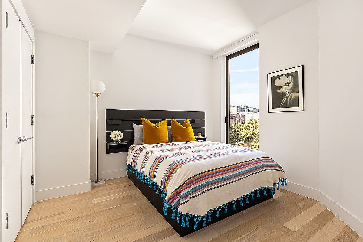 This is a bright and modern bedroom featuring a bed with a black headboard and a colorful striped blanket with tassels. The room has light wood floors, white walls, and a large window that lets in natural light. A black-framed artwork hangs on the wall, and a floor lamp stands near the bed, creating a cozy and inviting atmosphere.