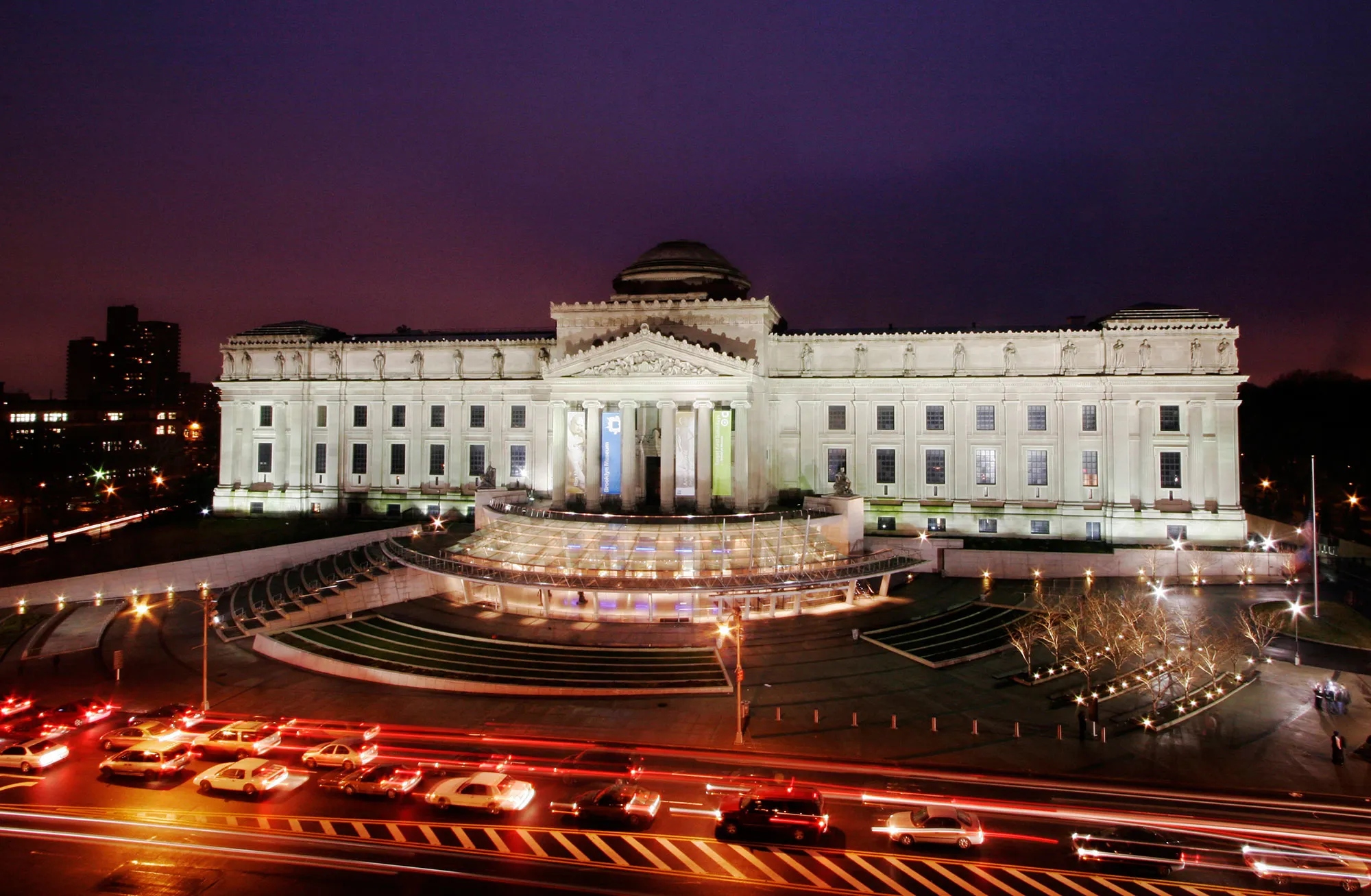 This image features the Brooklyn Museum at night, showcasing an impressive Beaux-Arts architectural style. The building is illuminated, highlighting its grand columns and intricate facade details. Light streaks from passing vehicles add a dynamic element to the scene, creating a vibrant backdrop for the museum's facade.